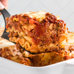 A slice of lasagna bolognese is being lifted from the baking dish.