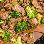A wooden stirring spoon is placed in a skillet filled with beef and broccoli.