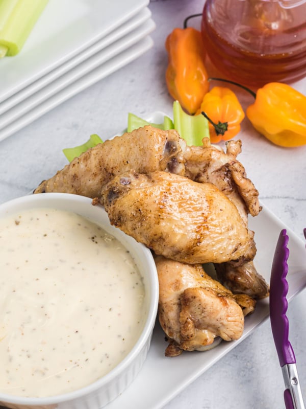 Several wings are placed on a white plate next to a bowl of white dipping sauce.