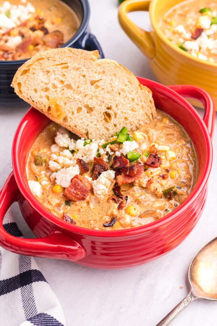 A small red bowl is filled with chowder and topped with a small slice of bread.