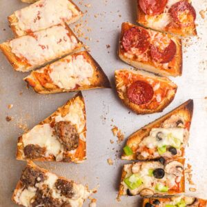 Sliced portions of french bread pizzas are all topped with different toppings on a baking sheet.