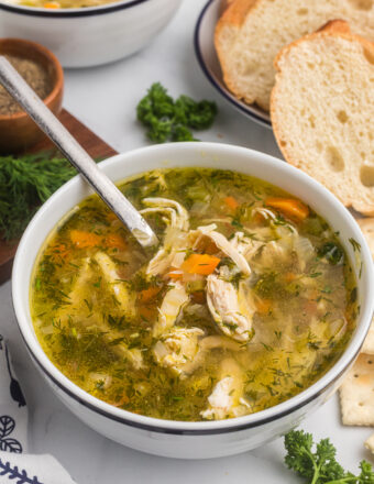 Chicken soup in a bowl with a spoon and slices of crusty bread in the background.