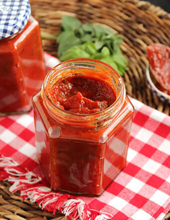 Homemade pizza sauce in a jar on a white and red checkered towel with basil in the background.