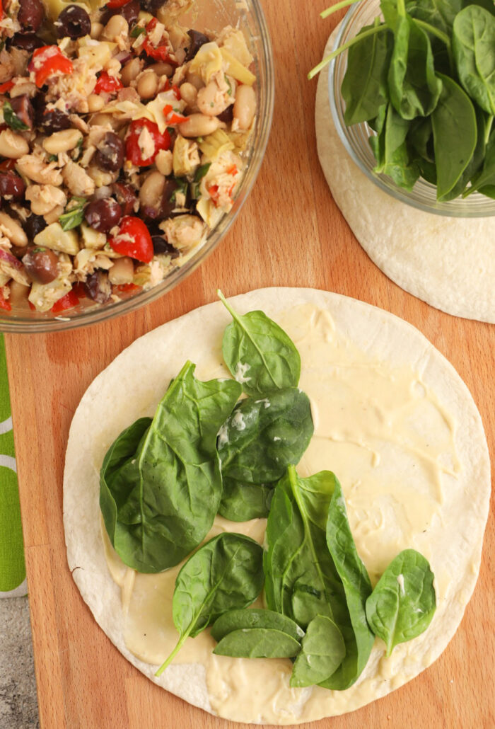 Overhead view of 2 tortillas with spinach leaves on top next to bowl of Mediterranean tuna salad