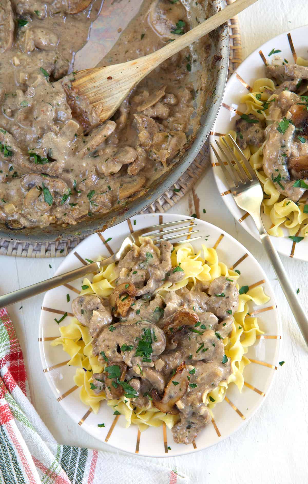 A plate of egg noodles is topped with beef stroganoff and placed next to a full skillet.