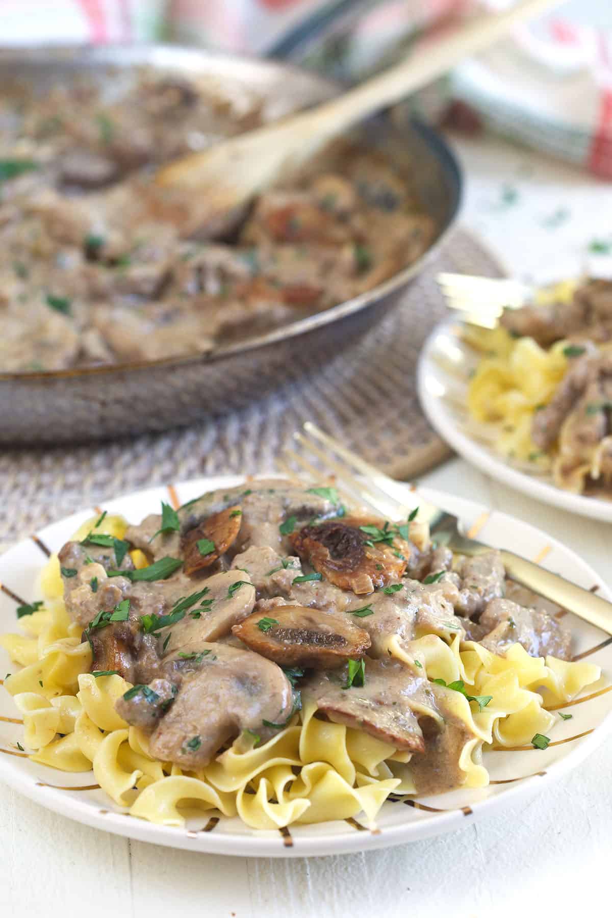 A plate of egg noodles and stroganoff is placed next to a skillet with a wooden spoon.