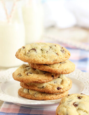 Five chocolate chip cookies stacked on a white plate placed on a blue, pink and white plaid napkin.