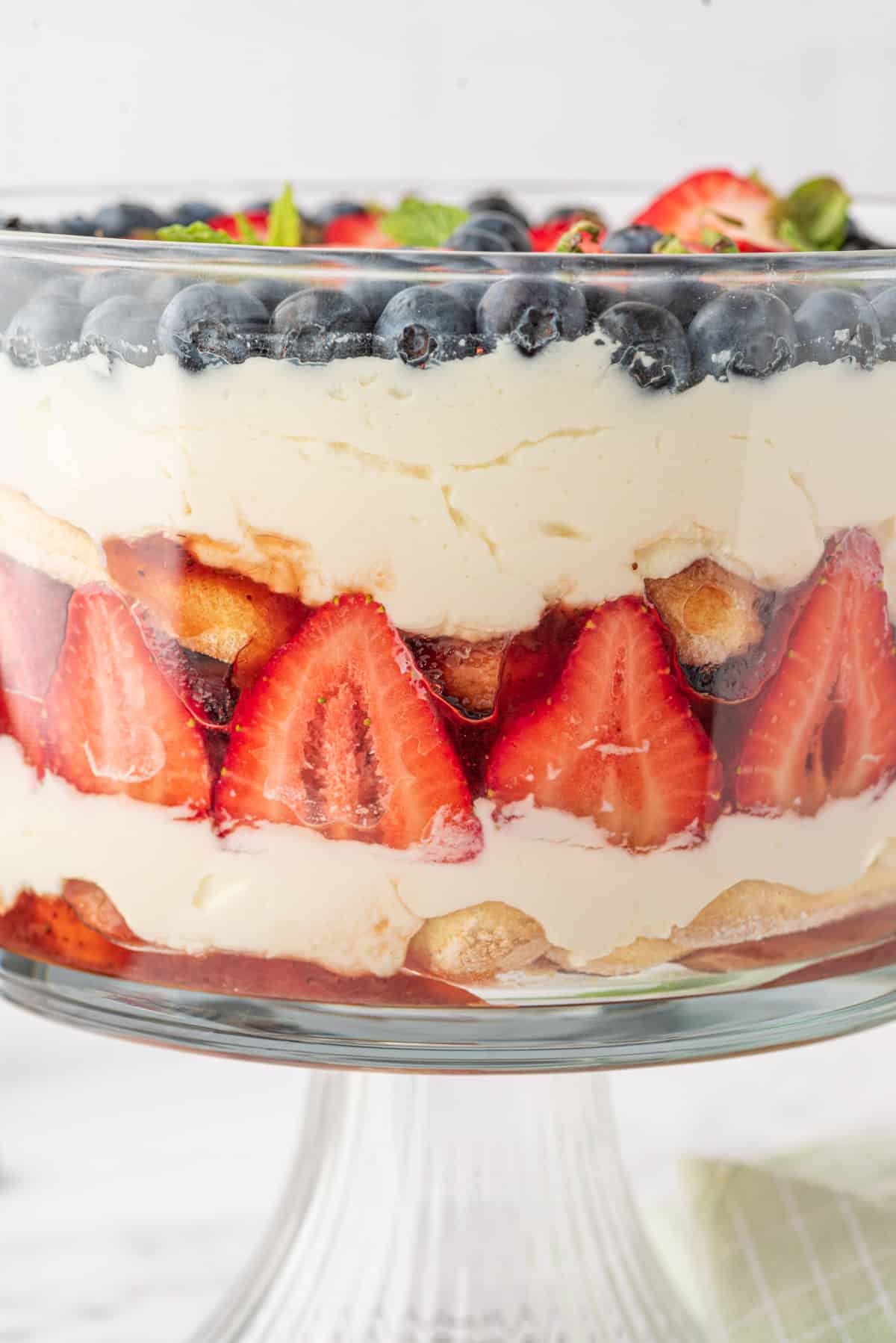 Berries, cream, and ladyfinegrs are layered in a glass trifle dish.