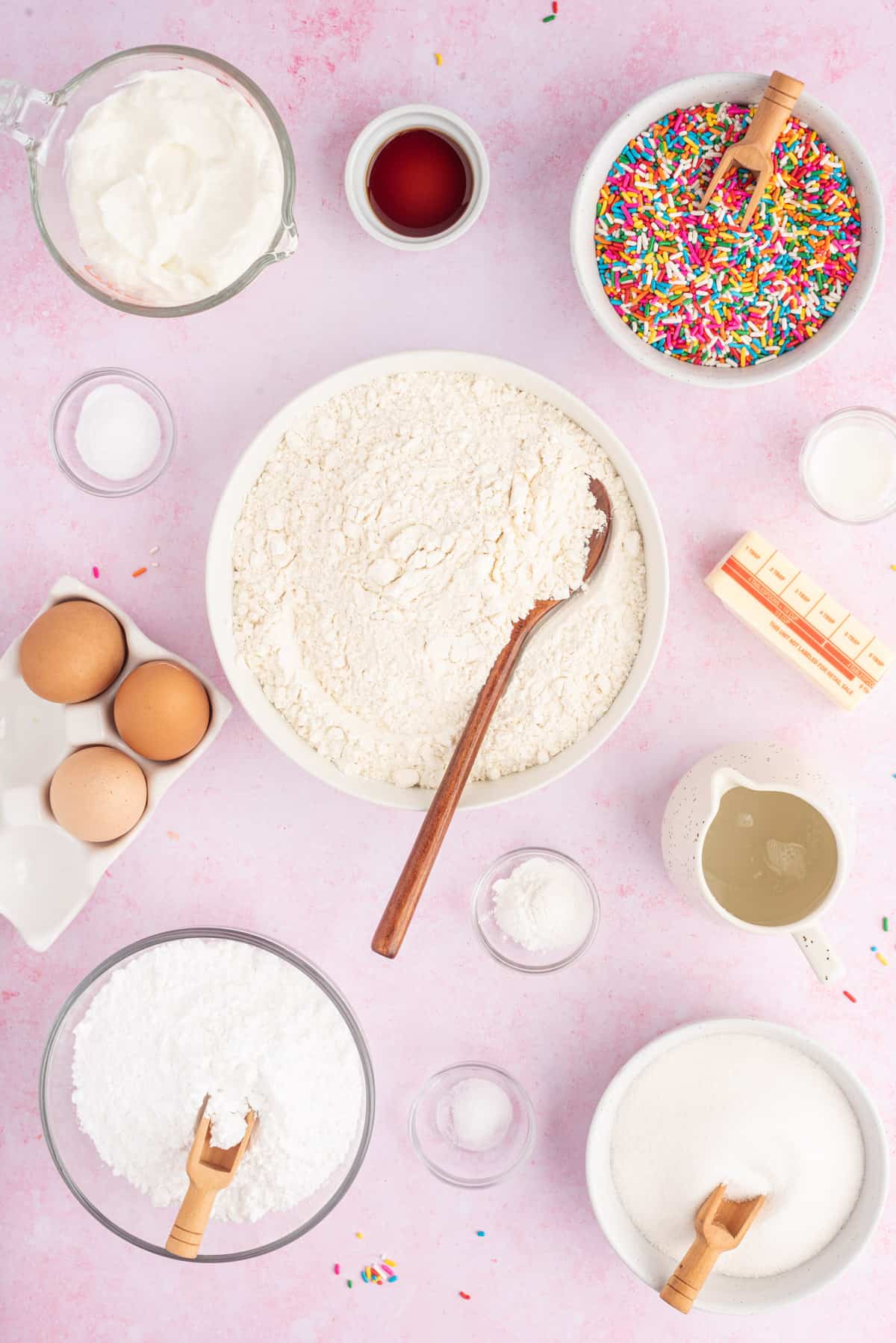 The ingredients for funfetti muffins are placed on a pink surface.