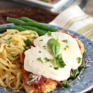 Chicken Parmesan on a blue plate with a side of spaghetti and garnished with tiny basil leaves.