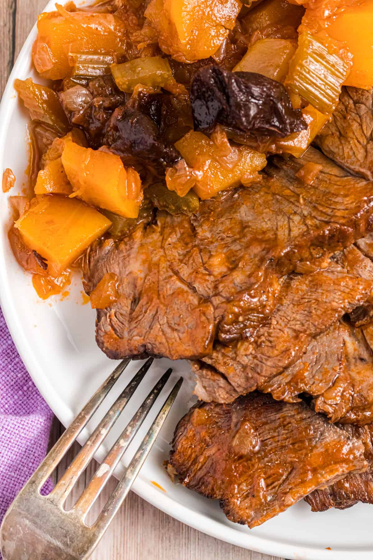 Pot roast, butternut squash and plums are plated on a round white plate with a metal fork.