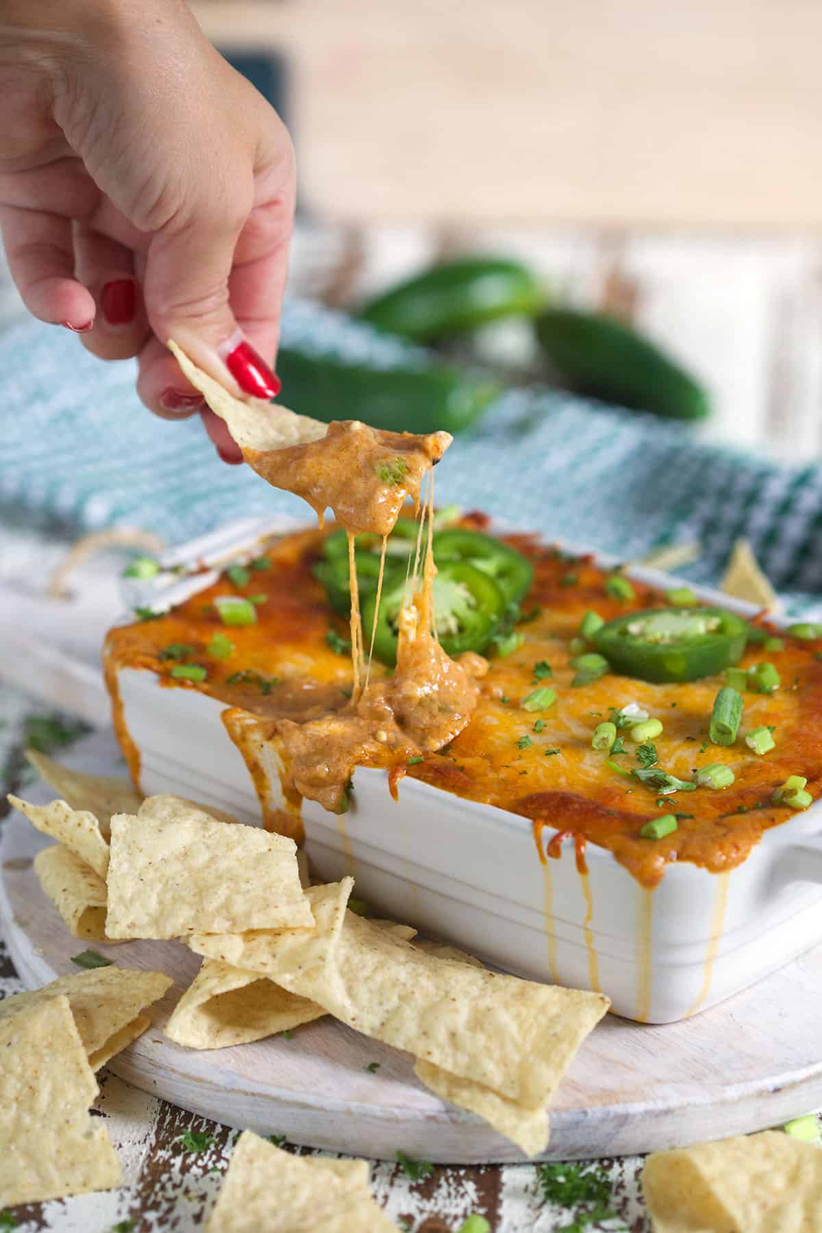 Chili Cheese Dip in a white baking dish with tortilla chips on the side and a hand holding a chip with a cheese pull to the dip.