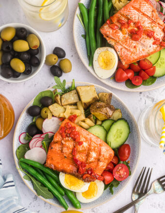 Two dinner plates with salmon Nicoise salad on them with a bowl of olives and two forks.