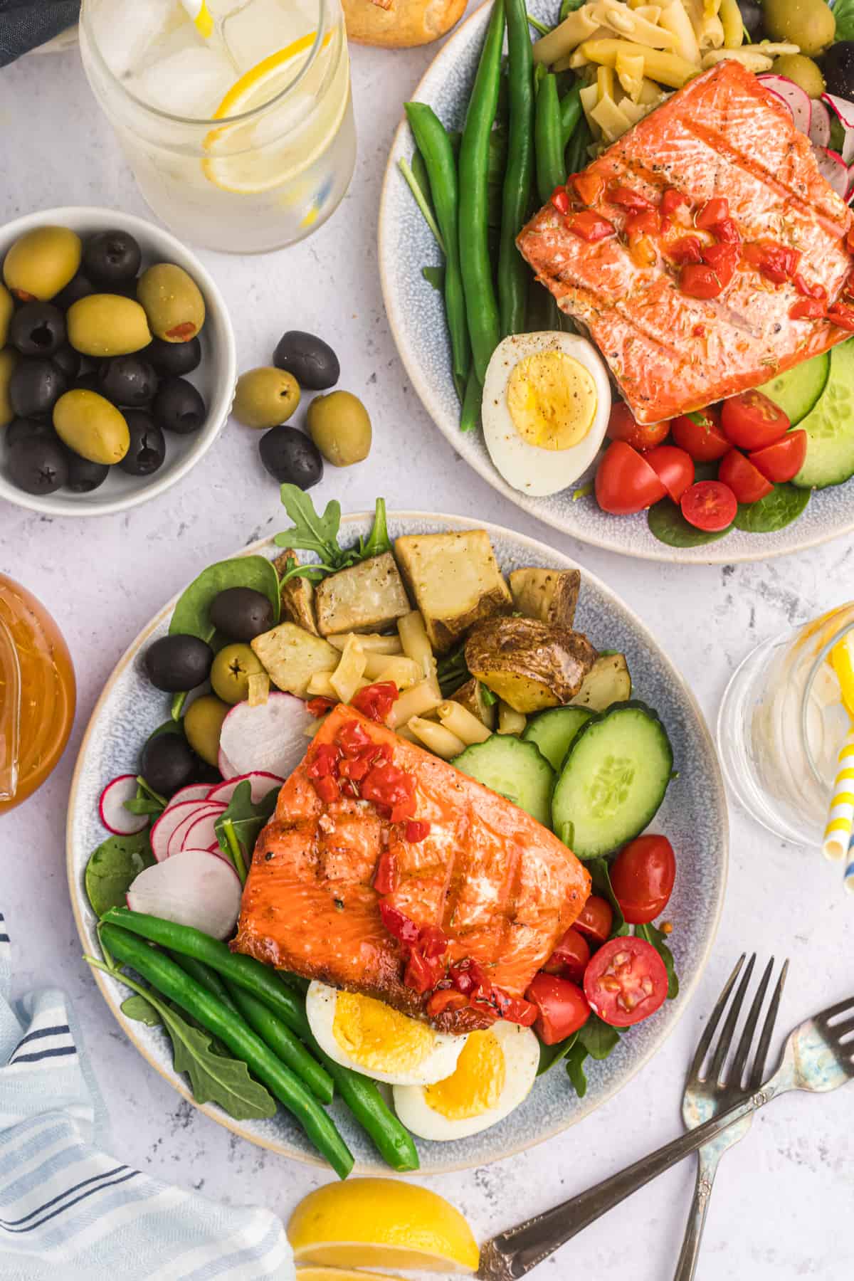 Two dinner plates with salmon Nicoise salad on them with a bowl of olives and two forks.