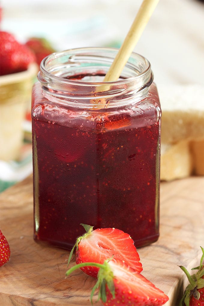 Strawberry jam in a jelly jar with a wooden spoon and two slices strawberries in front on a wooden board.