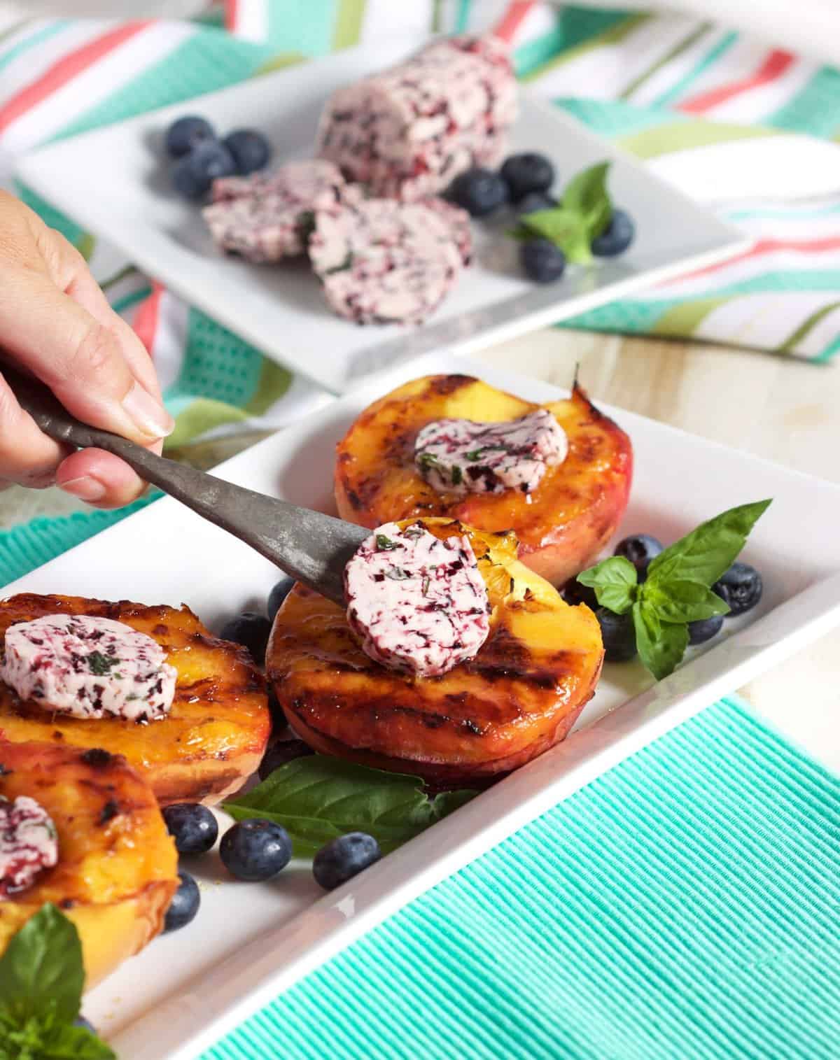 Grilled peaches on a white platter with blueberry compound butter being placed on top with a spreader.