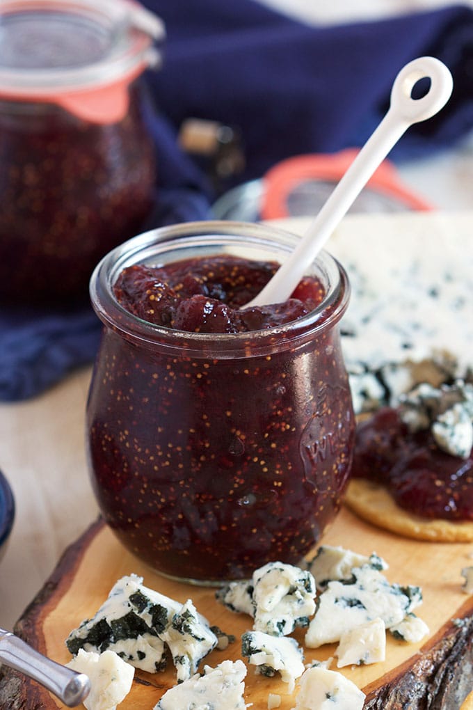 Fig jam in a tulip shaped jar with a white ceramic spoon on a wooden board with blue cheese.