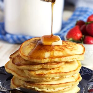 Pile of pancakes on a blue plate with a pat of butter on top and syrup being drizzled over it.