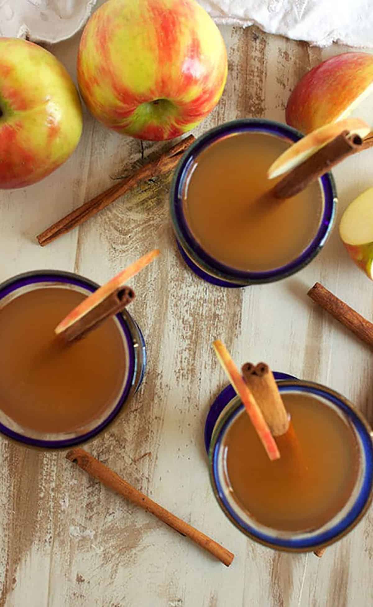 three glasses filled with apple cider with cinnamon sticks on a distressed wood background with apples.