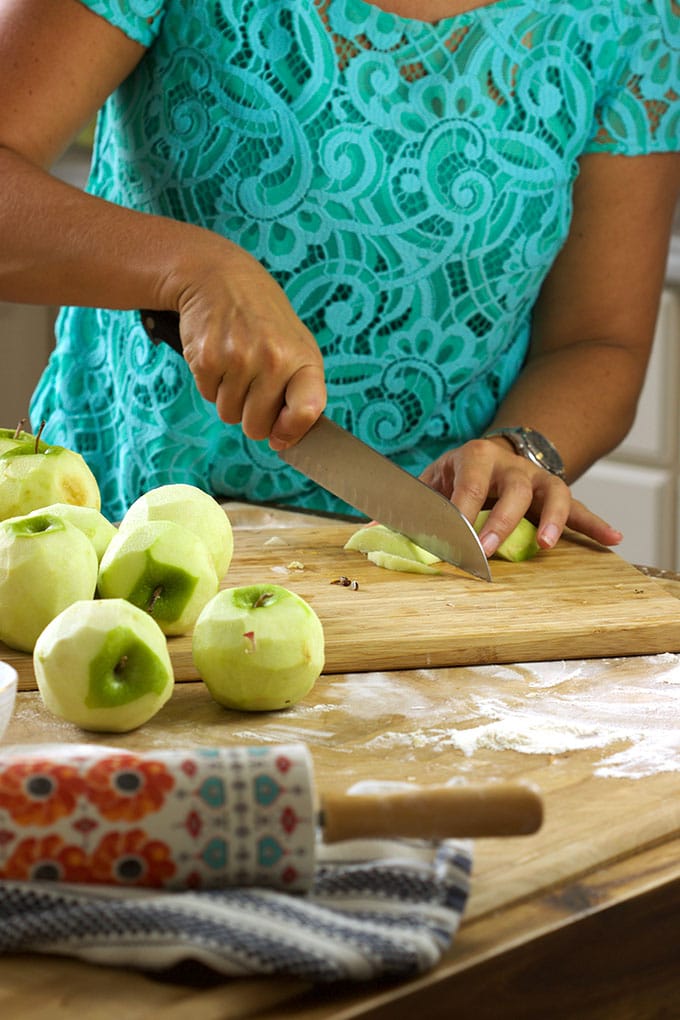 A women in a blue lace shirt is slicing several green apples on a cutting board. 