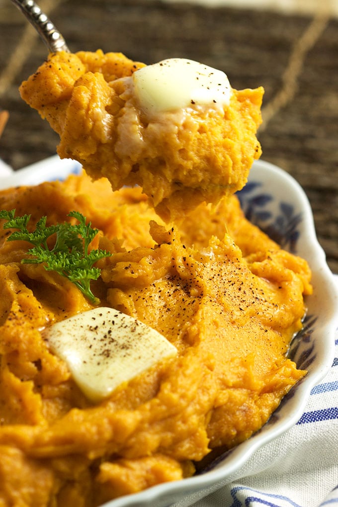 Serving spoon with mashed sweet potatoes on it topped with a pat of butter over a bowl of sweet potatoes.