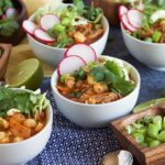White bowls with Pork Pozole Rojo and limes in a wooden square bowl.