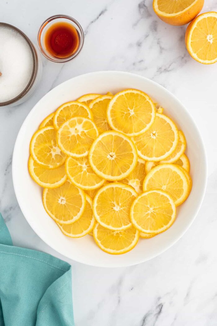 A white bowl full of orange slices is placed next to vanilla and sugar.