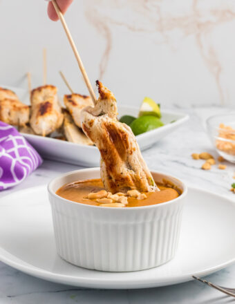 Grilled chicken on a skewer is being dipped into a cup of peanut sauce.