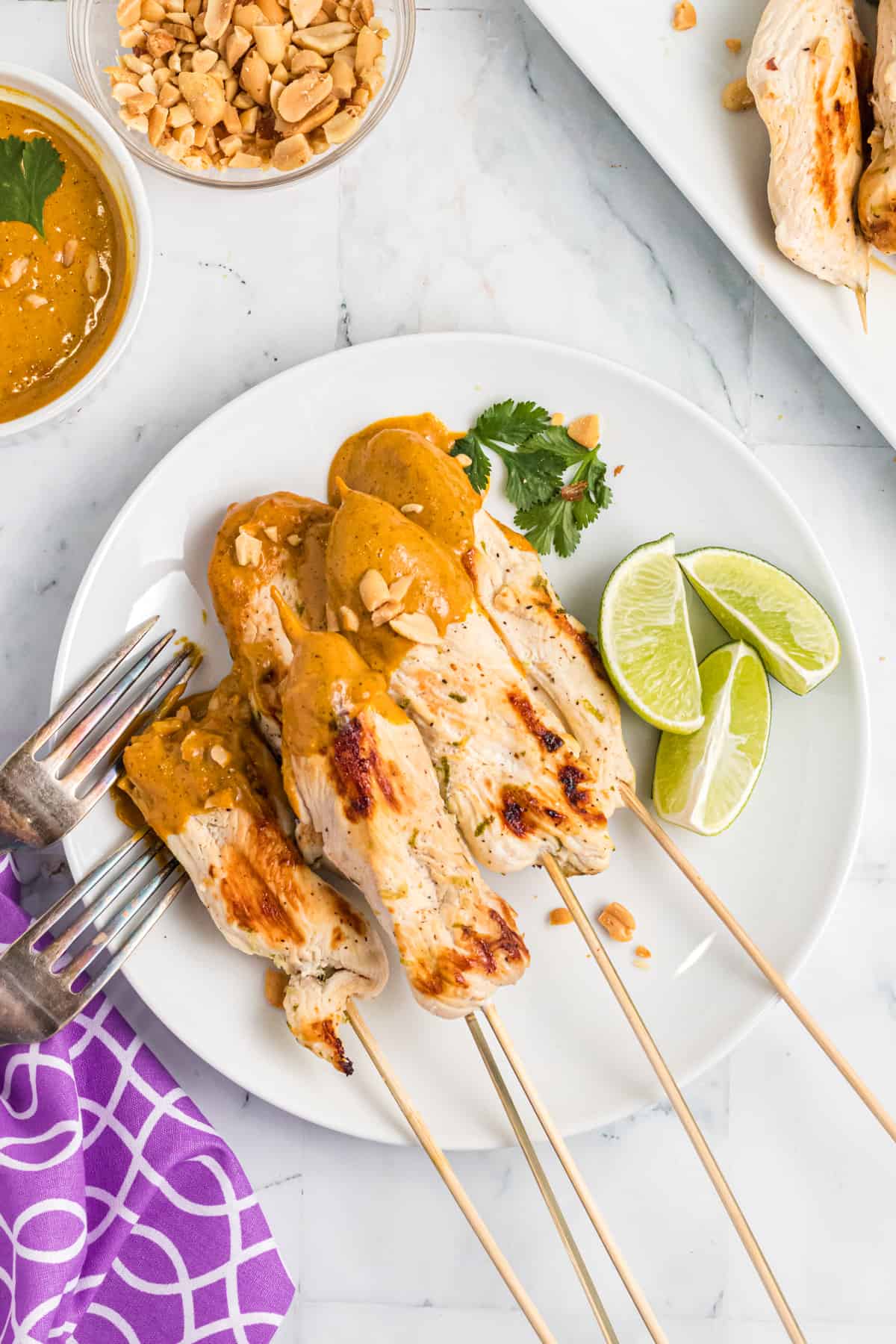 Chicken satay skewers on a white plate have been all dunked in peanut sauce and are placed next to fresh limes.