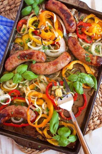 Sausage peppers and onions on a sheet pan with basil on a white board.