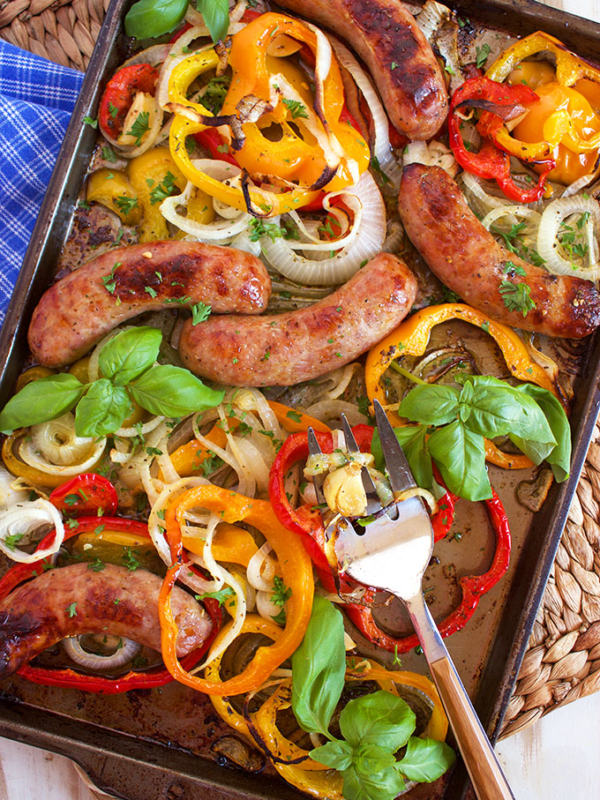 Sausage peppers and onions on a sheet pan with basil on a white board.