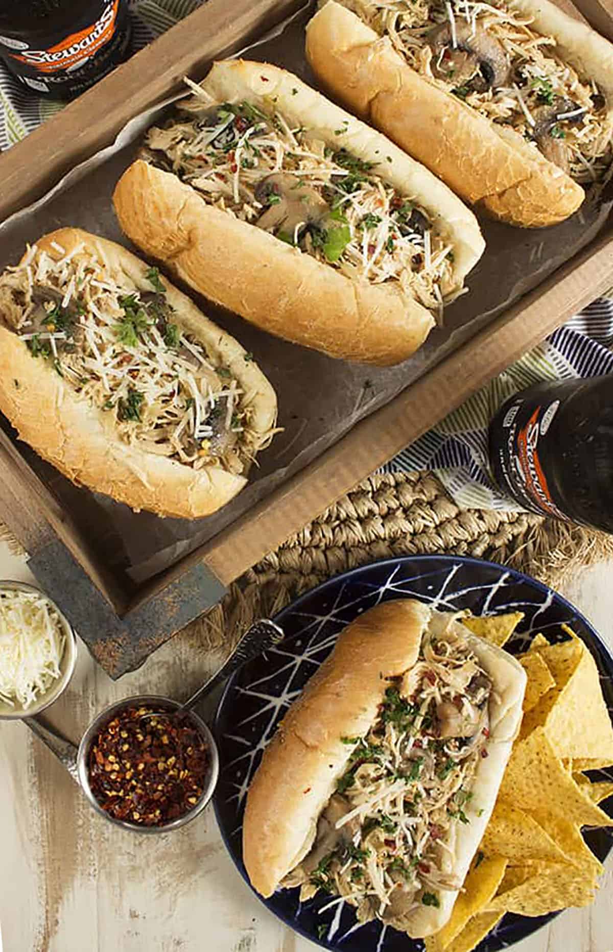 Three chicken cheesesteaks in a serving tray with one sandwich on a plate with chips.