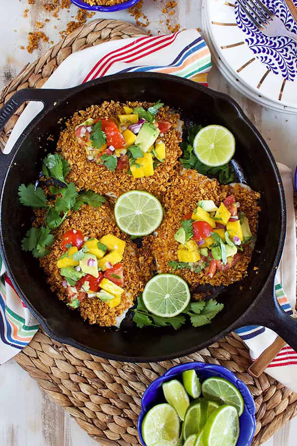 Tortilla crusted tilapia in a black cast iron skillet with sliced limes in a blue bowl.