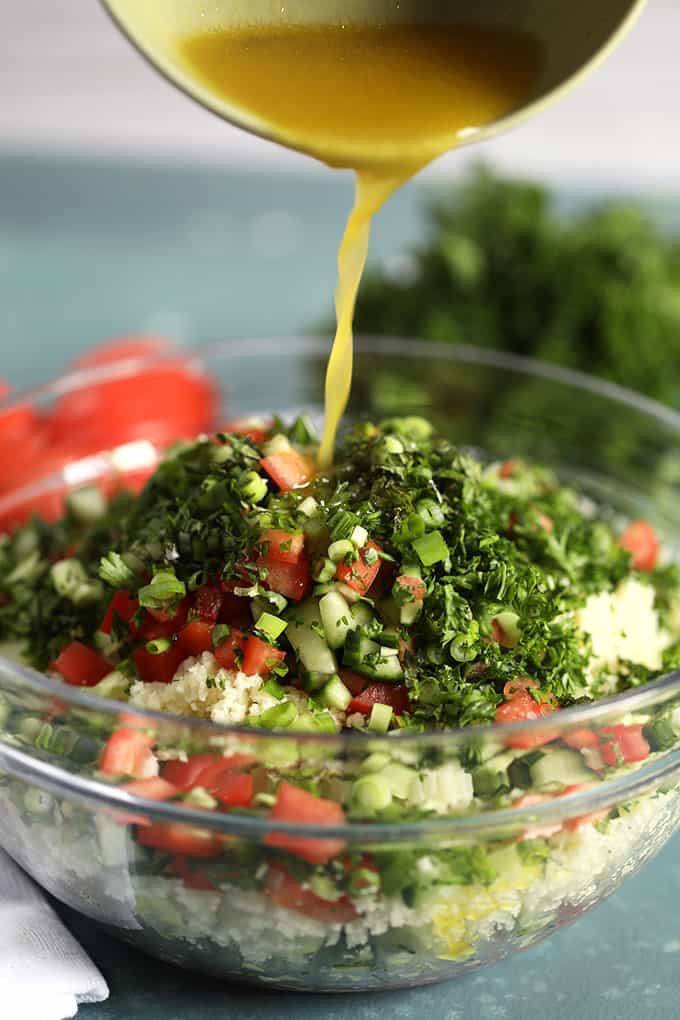 Dressing being poured over Cauliflower Rice Tabbouleh salad in a glass bowl on a blue background from TheSuburbanSoapbox.com