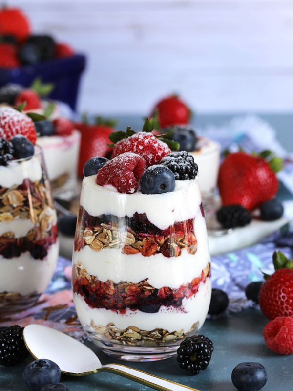 Layered cheesecake parfait with berries and muesli in a wine glass on a blue background from TheSuburbanSoapbox.com