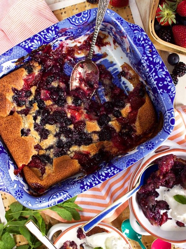 Overhead view of Berry Cobbler in a blue and white baking dish with two white dishes with spoons and whipped cream from TheSuburbansoapbox.com