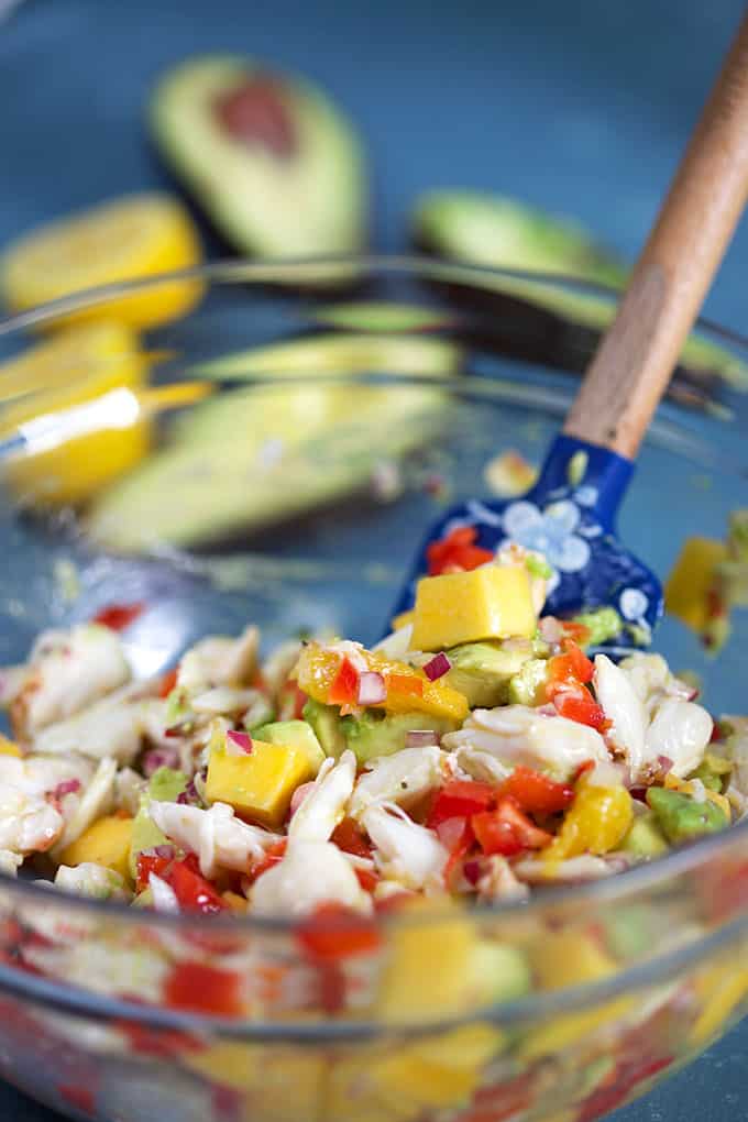 Mango Crab Avocado Salad in a glass bowl with a blue spatula stirring the salad from TheSuburbansoapbox.com