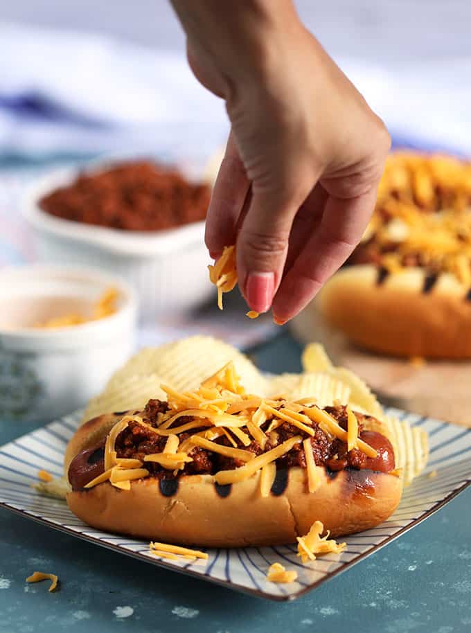 Hand placing shredded cheese on a chili cheese dog from TheSuburbanSoapbox.com