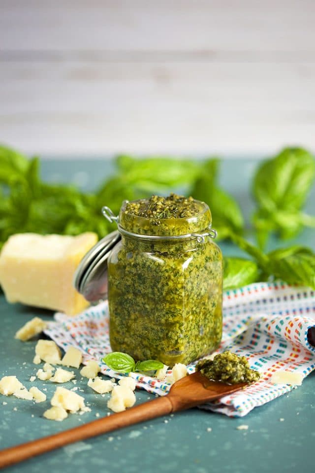 The Very Best Basil Pesto Sauce in a glass jar with a wooden spoon on a blue background with basil leaves in the background from TheSuburbanSoapbox.com