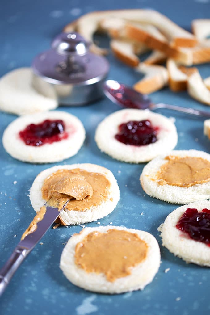 Peanut butter and jelly being spread on white bread rounds on a blue background from TheSuburbansoapbox.com
