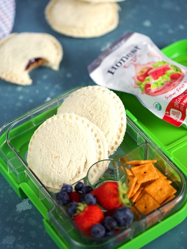 Homemade Peanut Butter and Jelly Uncrustables in a green lunchbox with fruit and crackers on a blue background from ThesuburbanSoapbox.com