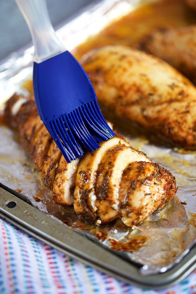 Sliced baked chicken breast being basted with a blue silicone basting brush on a foil lined baking sheet.