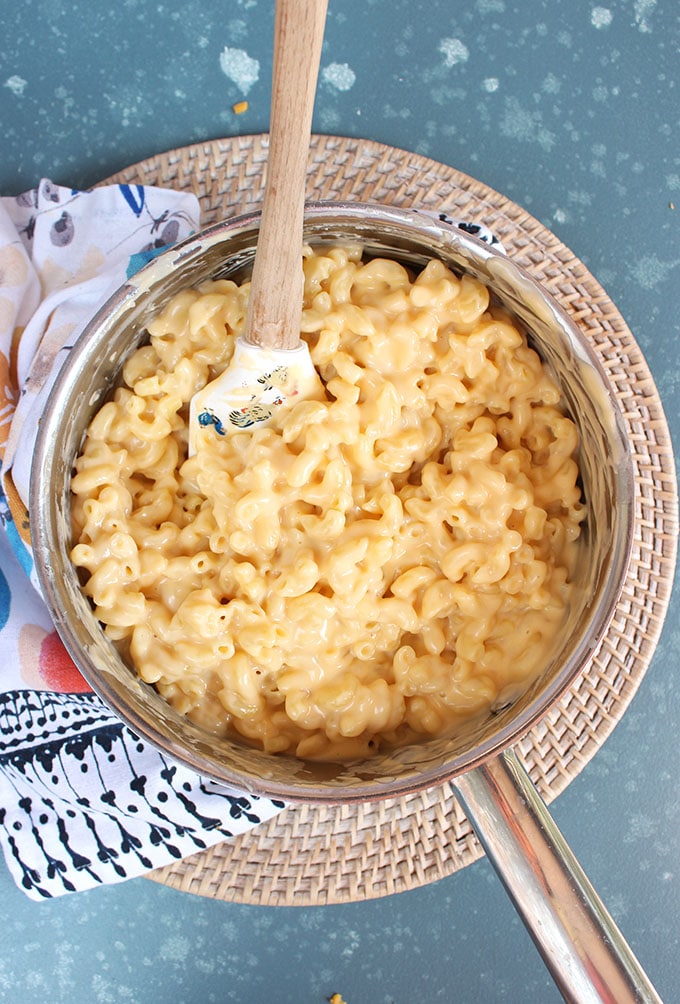 Finished Mac and cheese recipe in a silver saucepan with a wooden spatula on a blue background from TheSuburbansoapbox.com