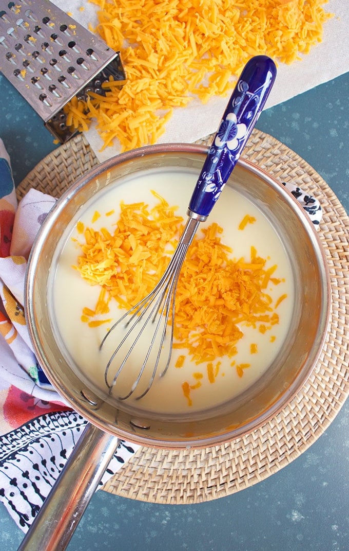 Cheddar Cheese being added to milk mixture for easy Mac and cheese recipe in a silver saucepan with a blue floral whisk from TheSuburbanSoapbox.com