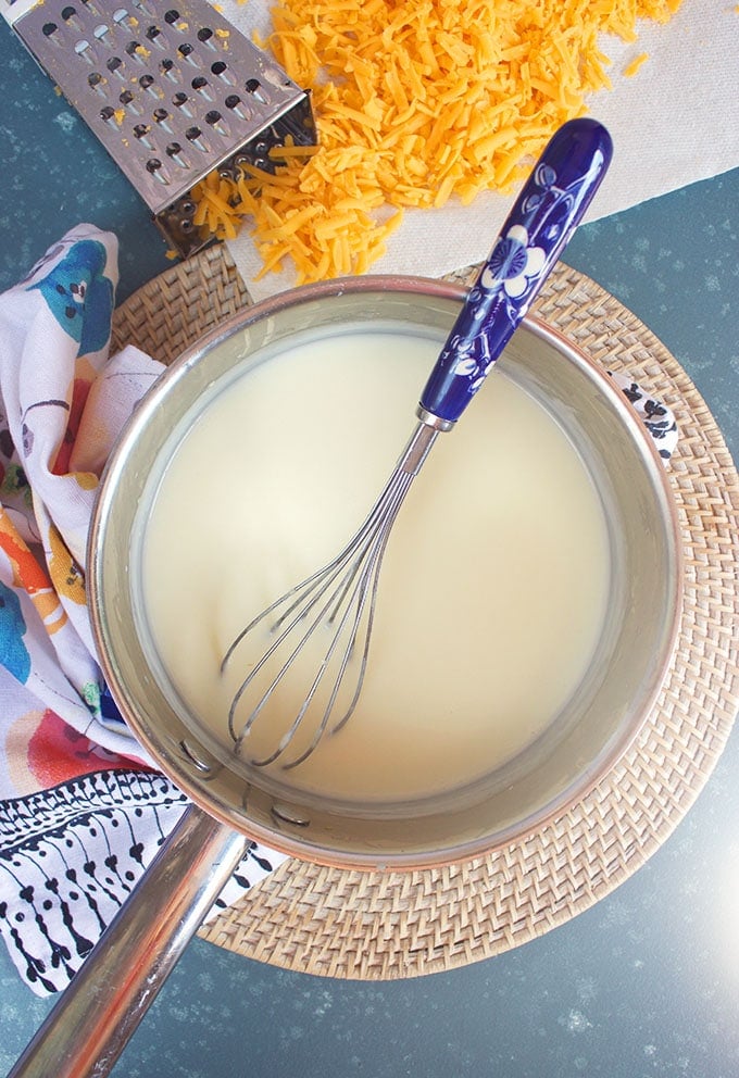 Milk mixture for Mac and cheese sauce in a silver saucepan with a blue whisk.