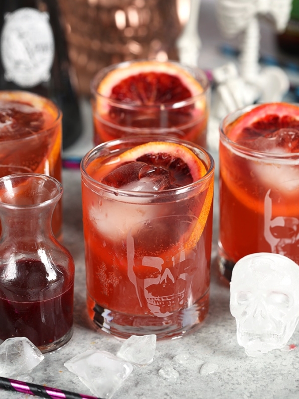 Far shot of four dark and stormy cocktails in a high ball glass with a carafe of blood orange juice and paper straws.