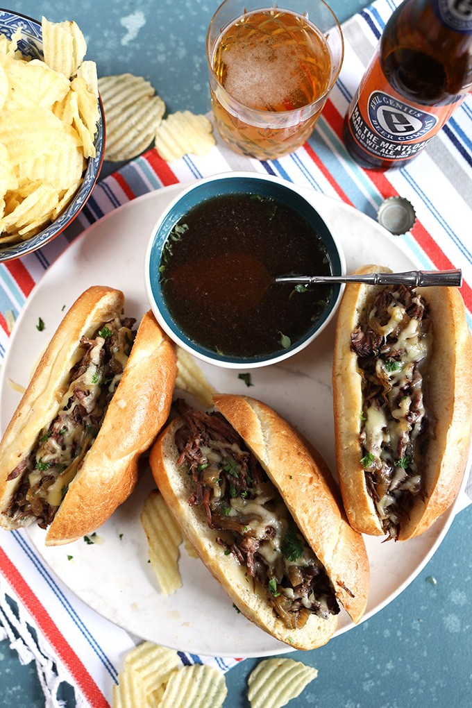 Overhead shot of three French dip sandwiches with a bowl of au jus on a white platter with a glass of beer and side bowl of chips.