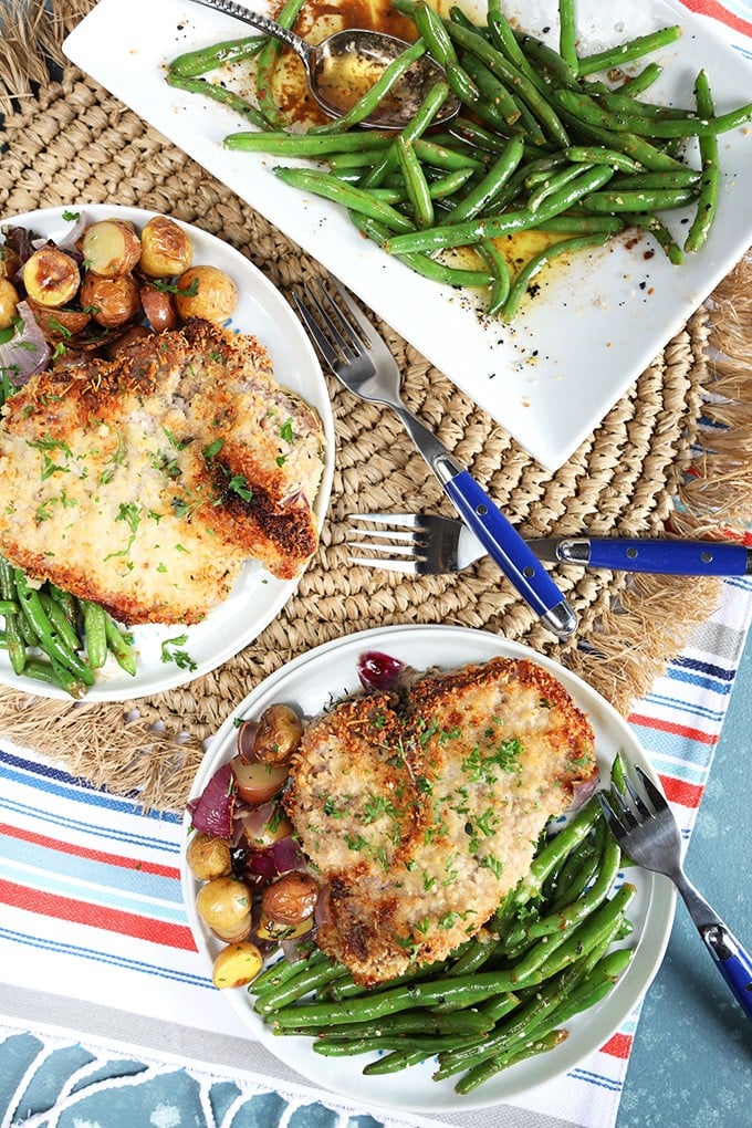 Overhead shot of two white plates with parmesan crusted baked pork chops and a serving of green beans on a white platter with brown butter sauce.