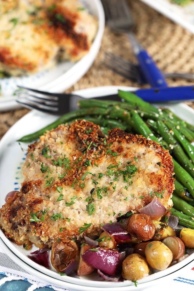 Close up shot of parmesan crusted baked pork chops with potatoes and green beans on a white plate.