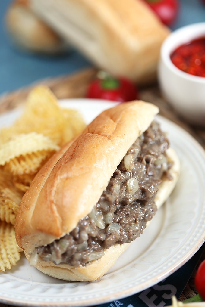 Philly cheesesteak sandwich on a white plate with chips and pepper sauce on the side.